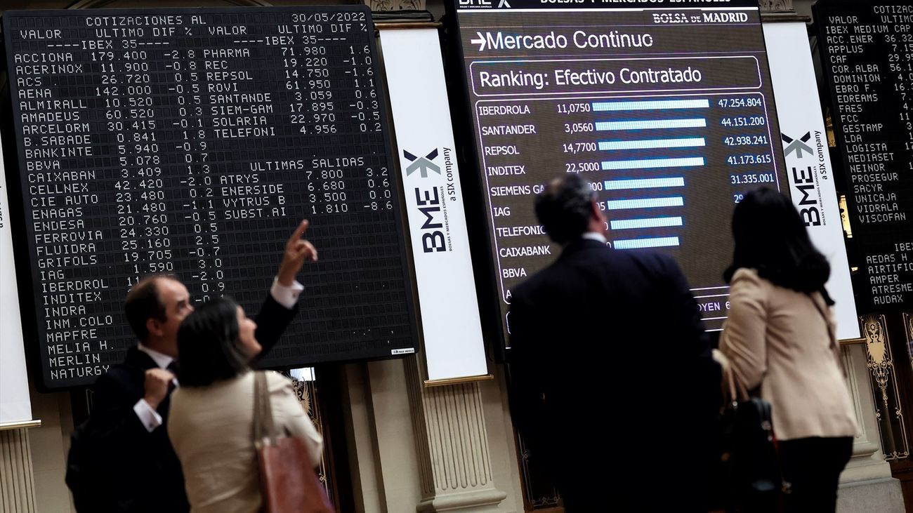 Cuatro personas frente a un panel de valores del Ibex en la Bolsa de Madrid